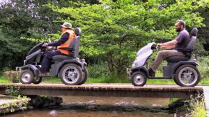 Side view of two tramper users on a wooden bridge running over some water at Sutton Park. Trees can be seen behind them.