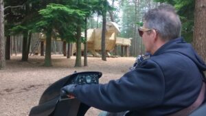Man in tramper on path at Moors Valley Country Park looks at large wooden play structure of bird.