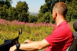 Man in Tramper exploring National Trust Killerton. In the distance are tall hot pink flowers. Trees and rolling hills can be seen in the distance.