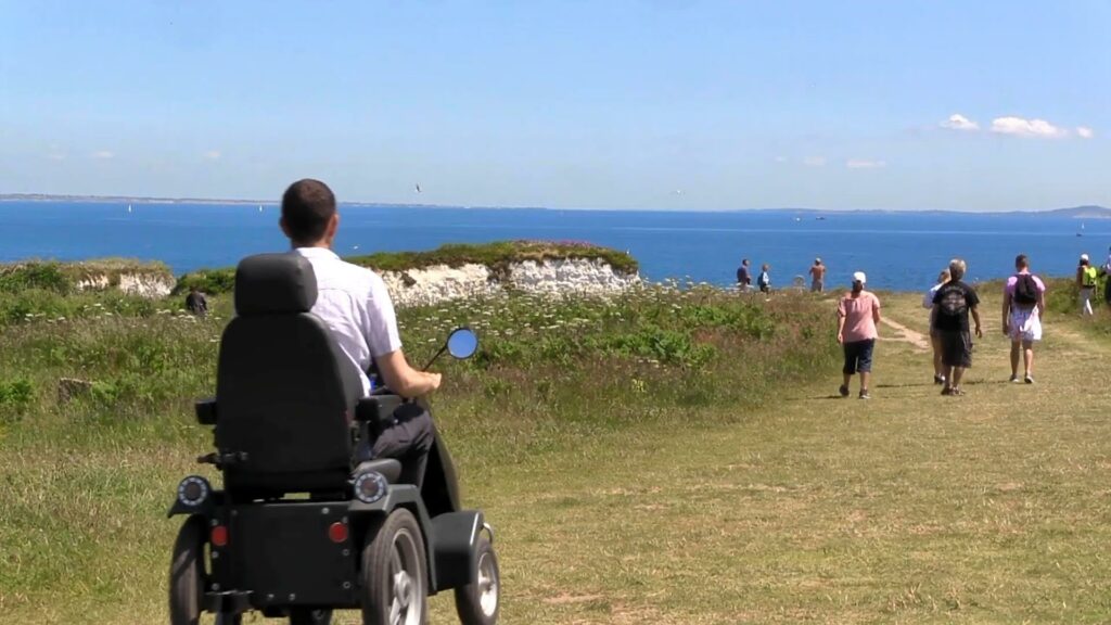 Man in tramper driving down a path towards a coast view at National Trust Studland. Sailboats and flying birds can be seen in the horizon. There are also other walkers on the path to the sea. It's a bright blue summers day, so everyone is dressed for the warm weather!