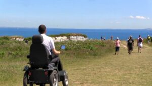 Man in tramper driving down a path towards a coast view at National Trust Studland. Sailboats and flying birds can be seen in the horizon. There are also other walkers on the path to the sea. It's a bright blue summers day, so everyone is dressed for the warm weather!