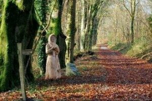 Tree-lined path at Buckland Abbey. The path is covered in fallen leaves. A statue wearing a cloak can be seen in the distance.