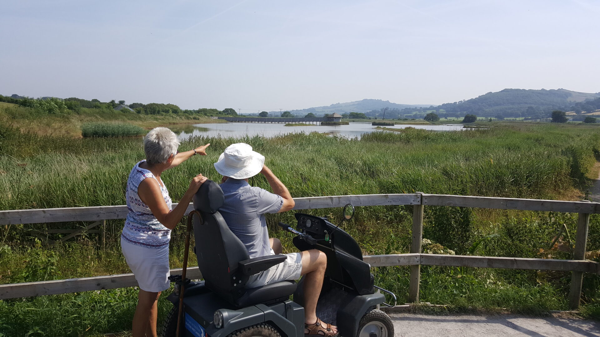 2 visitors (one sat in an all terrain scooter, one standing) look out across a wide view at Seaton Wetlands nature reserve