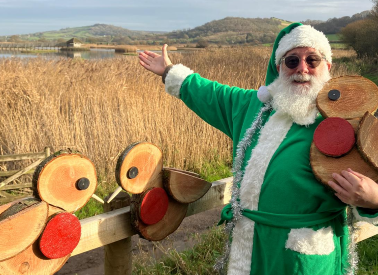 [Man wearing green father Christmas costume showing off some wooden Robins on a trail.]