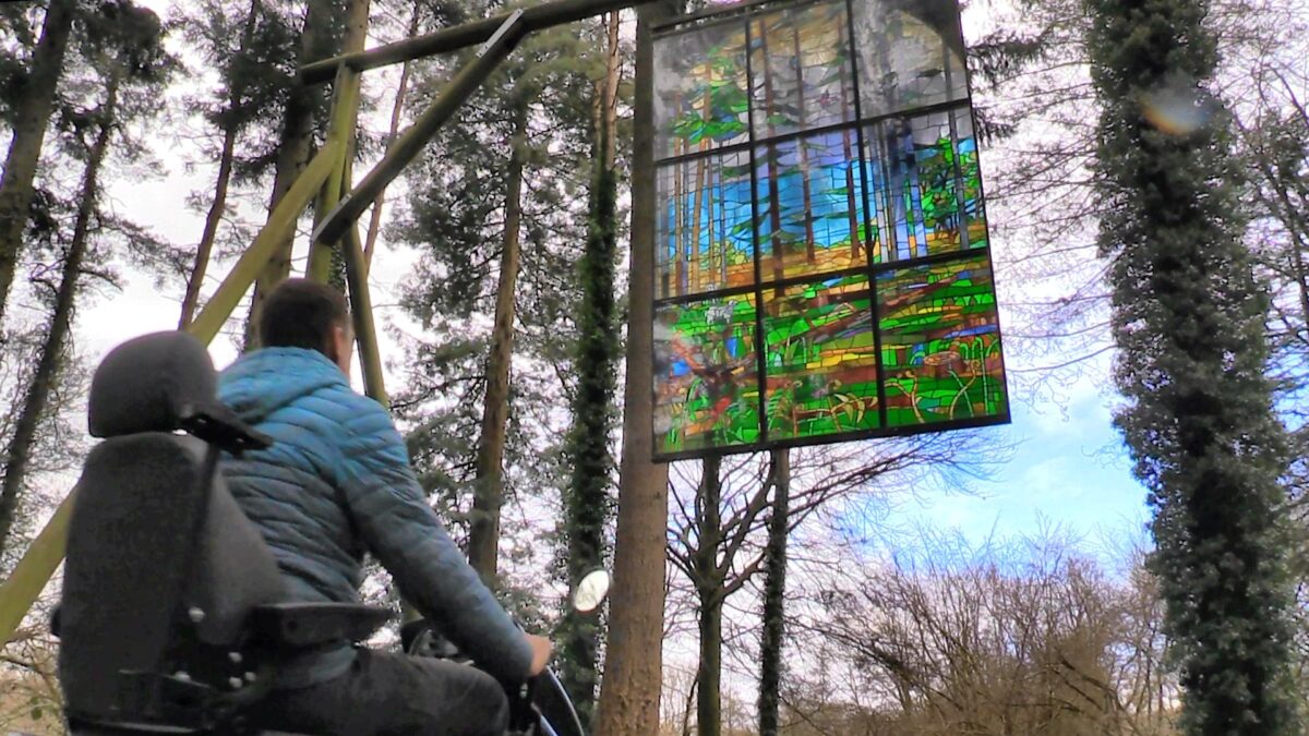 Visitor looks up from an all terrain mobility scooter to a stained glass window hanging from trees in a forest.