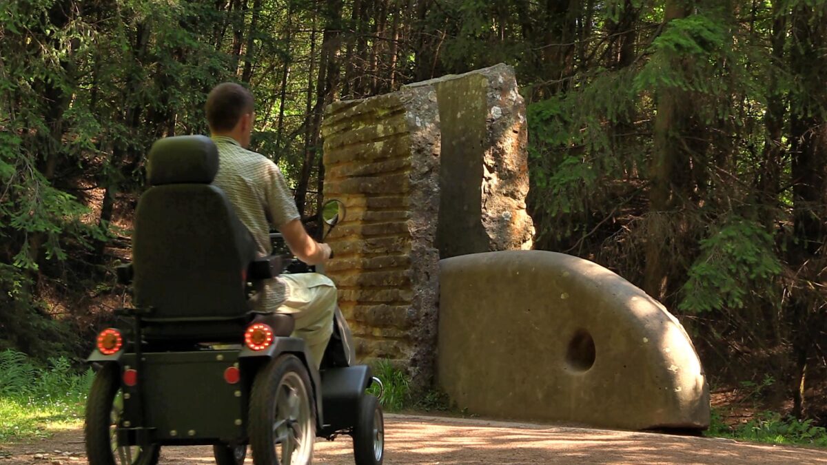 All terrain mobility scooter approaches a sculpture set within a forest