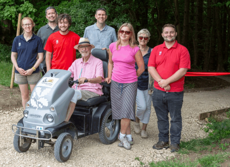 Mike, Loxie, Val, Neil, and the National Trust team stand in front of a red ribbon. Mike is on a Countryside Mobility Tramper