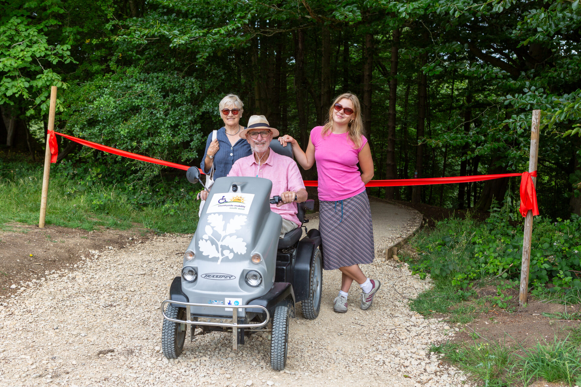 Loxie and Val stand either side of Mike, who is on a Tramper. They are in front of a red ribbon. They are smiling