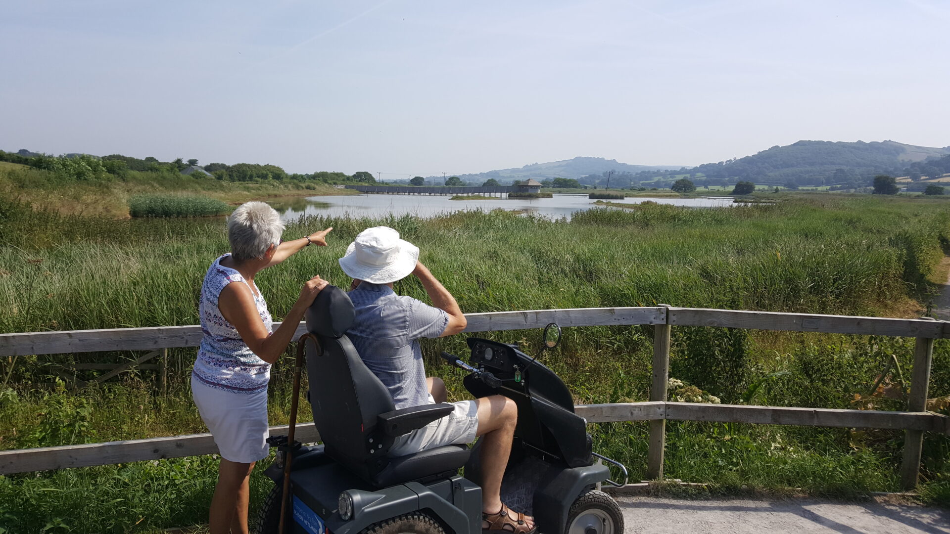 2 visitors (one sat in an all terrain scooter, one standing) look out across a wide view at Seaton Wetlands nature reserve 
