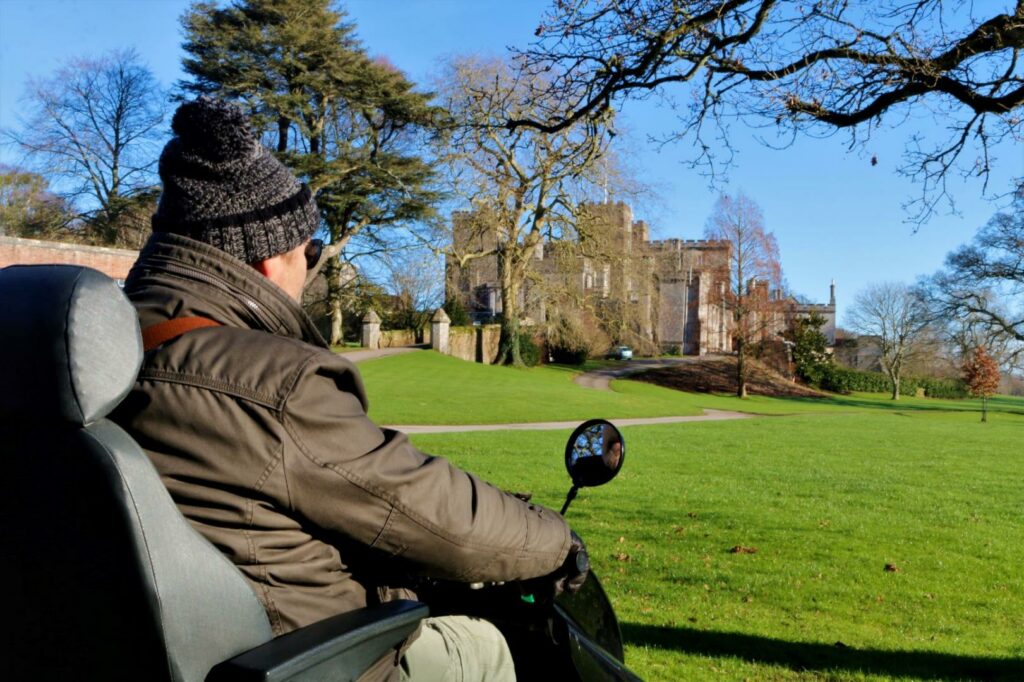 Man in Tramper on path looking out at Powderham Castle. The sky is bright blue, the man wears a woolly hat. 
