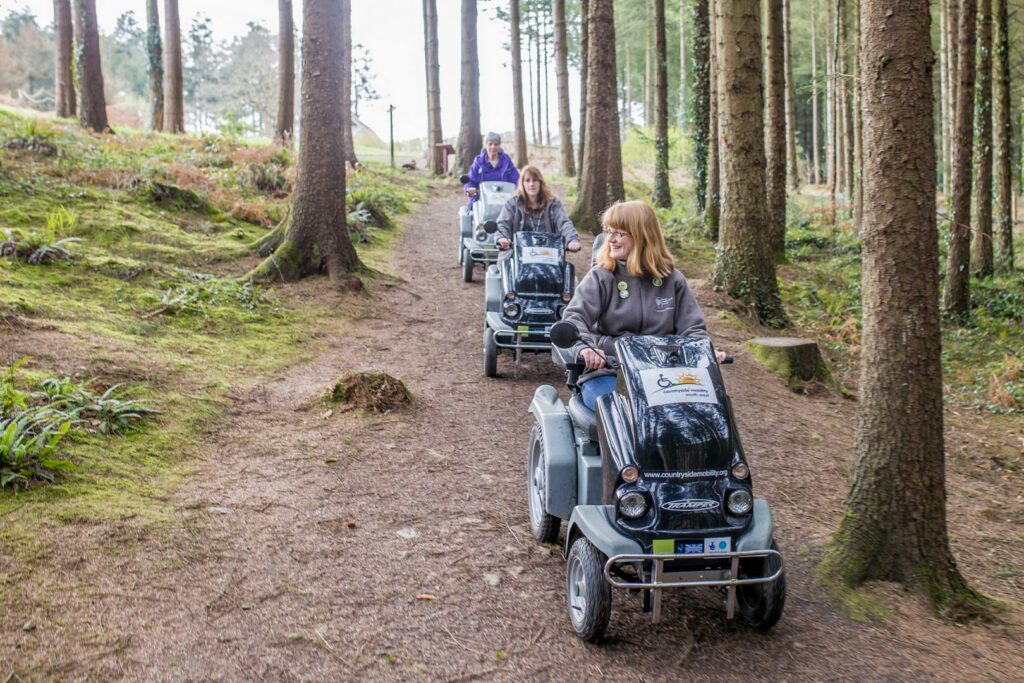 Three ladies on Trampers exploring the grounds of RHS Rosemoor. The path is lined by trees. 