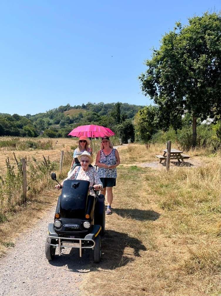 Eve smiling with her family while using the Tramper at Branscombe, with a view of a bench and trees behind her. Her family is holding a parasol.